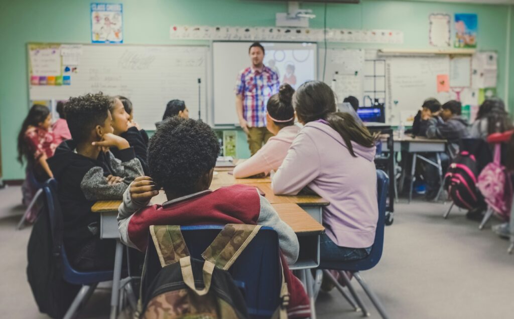 Male teacher in plaid shirt leads discussion in high school classroom with students arranged in desk clusters.